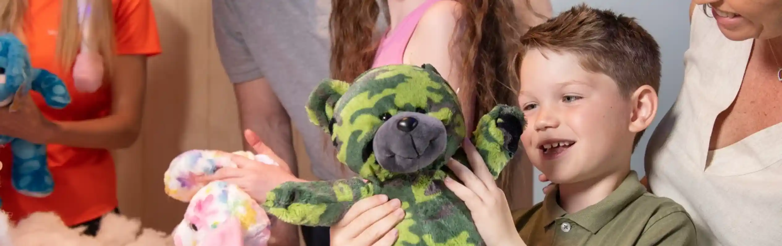 A smiling boy holds up a green camouflage teddy bear he has just made during a Stuff a Bear activity. Family members and staff watch happily, surrounded by colourful plush toys and stuffing.