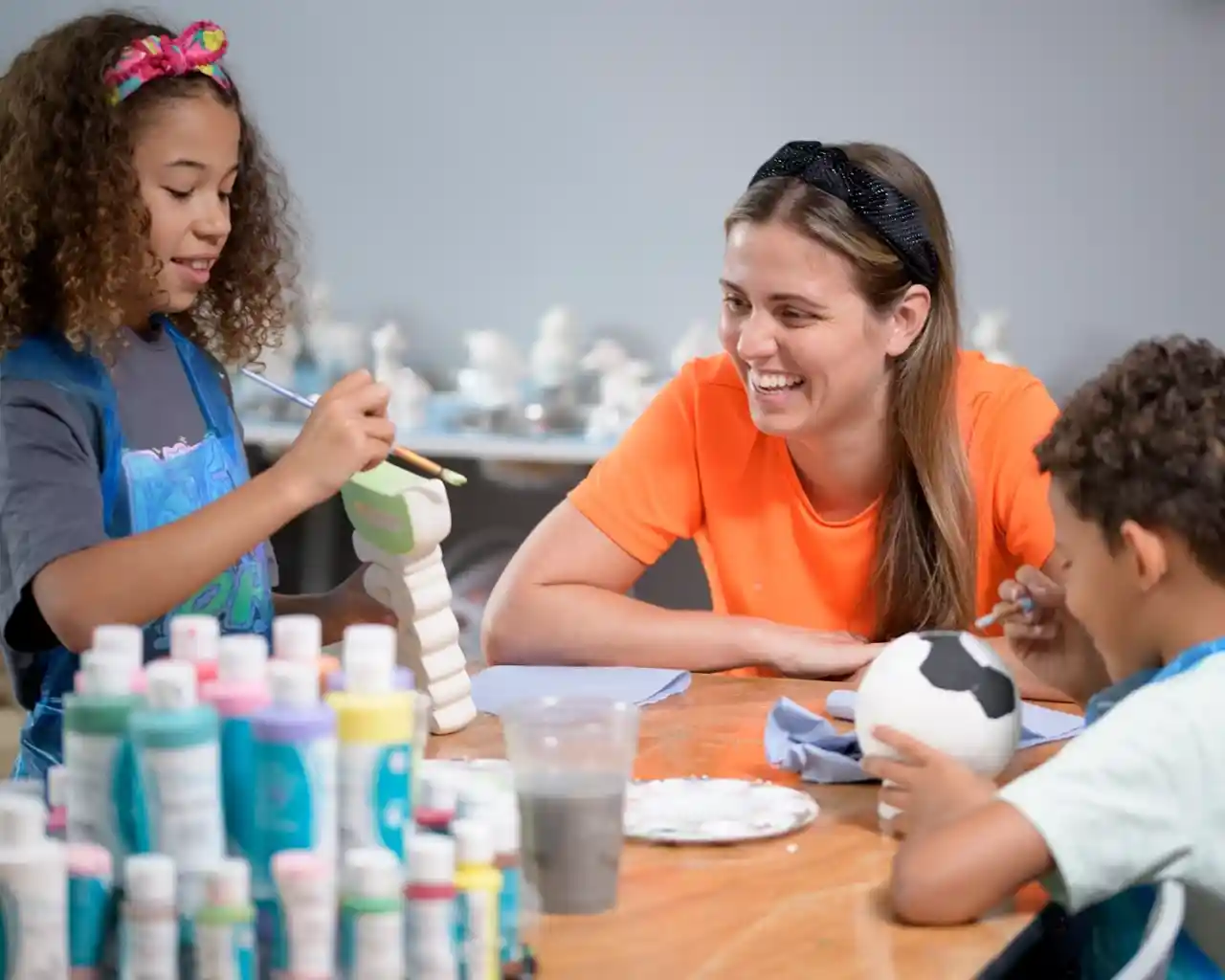 Two children and an adult enjoy a pottery painting activity together. One child is painting a ceramic figure, while the other paints a ball. The adult smiles warmly, creating a fun and creative atmosphere. Paints and brushes are spread across the table.