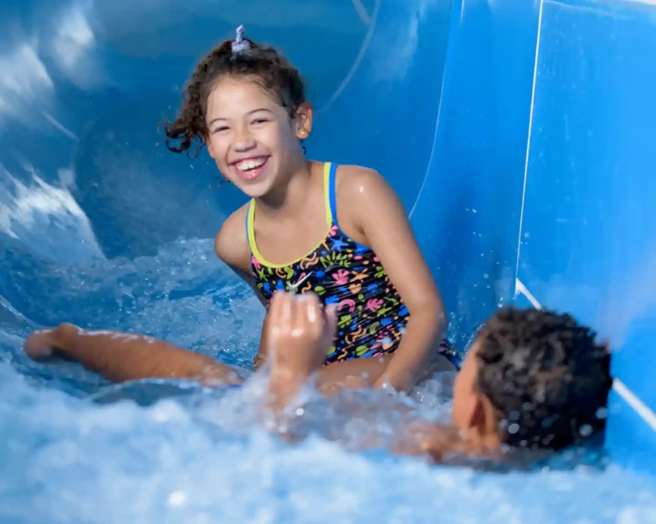 A young girl in a colourful swimsuit laughs as she rides down a water slide, while a boy splashes in the water below her. Both are enjoying a fun day at a water park.