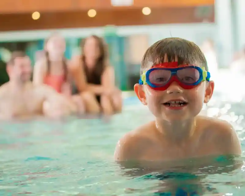 A smiling young boy wearing colorful goggles swims in a pool, with blurred adults enjoying the water in the background.