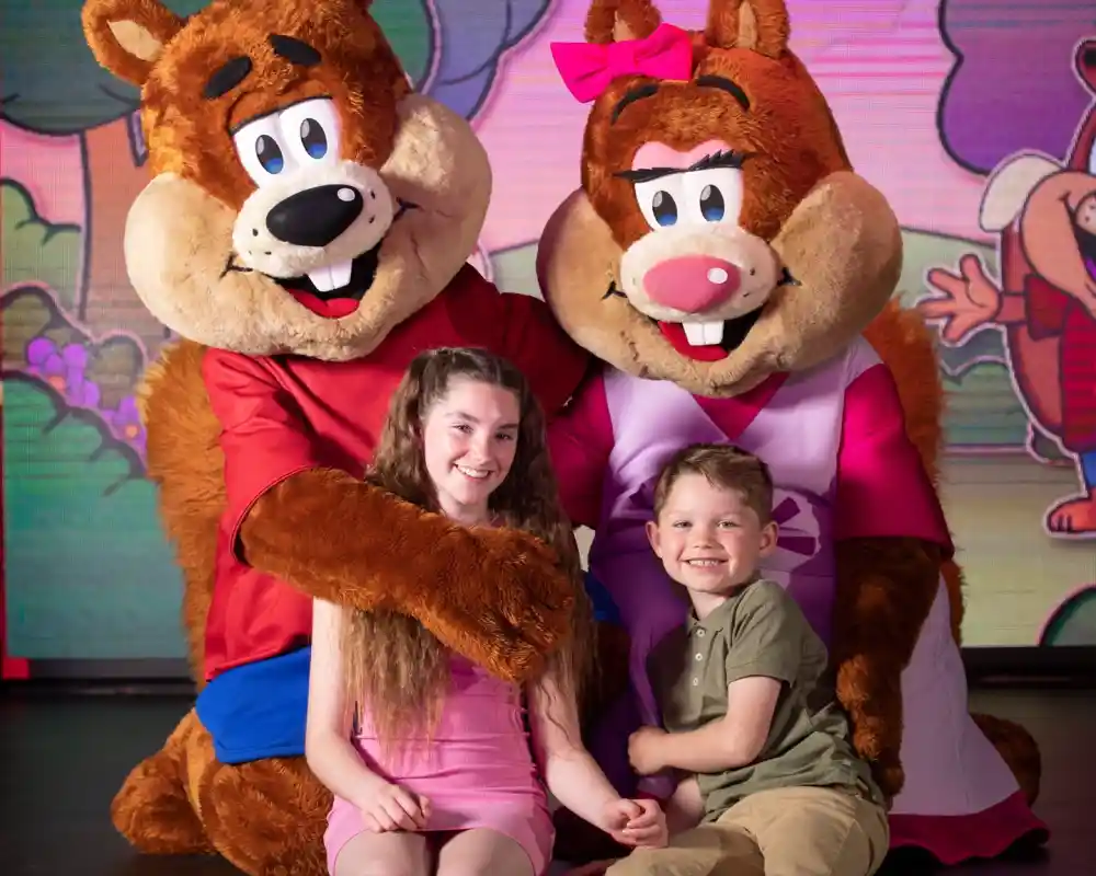 Two cartoon bear mascots with big smiles pose with a girl and a boy. The girl wears a pink dress, while the boy is in a light green shirt. They are in a colorful setting with cartoon backgrounds.