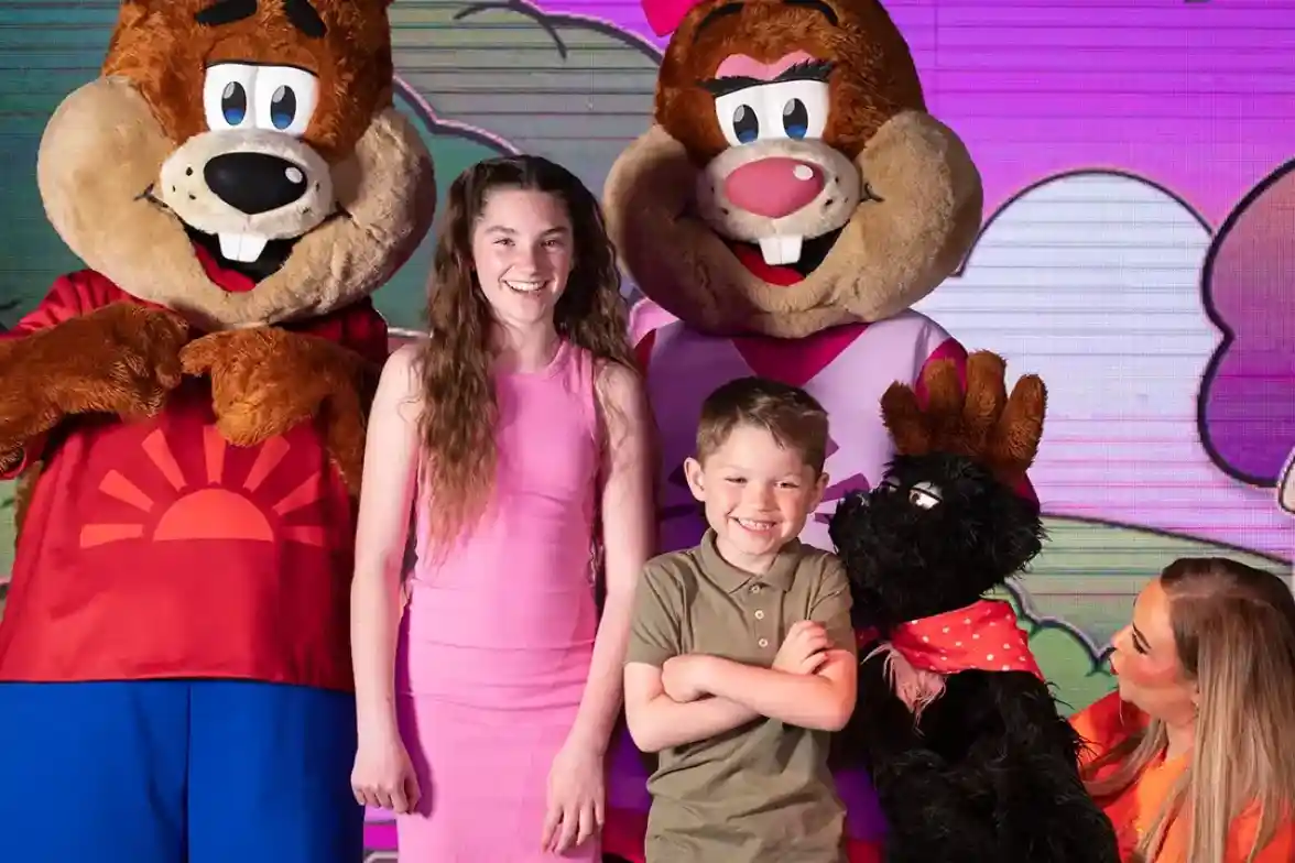 Two costumed characters resembling bears stand alongside a young boy and a girl. The boy crosses his arms while the girl smiles, wearing a pink dress. A black furry puppet with a red scarf is also present, and a colorful, cartoonish background adds vibrancy to the scene.