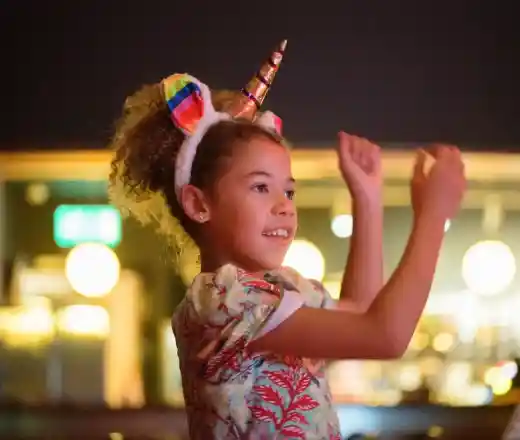 A young girl with curly hair wearing a rainbow-striped unicorn headband smiles and raises her arms in a celebratory pose. Soft lights create a warm, inviting atmosphere in the background.