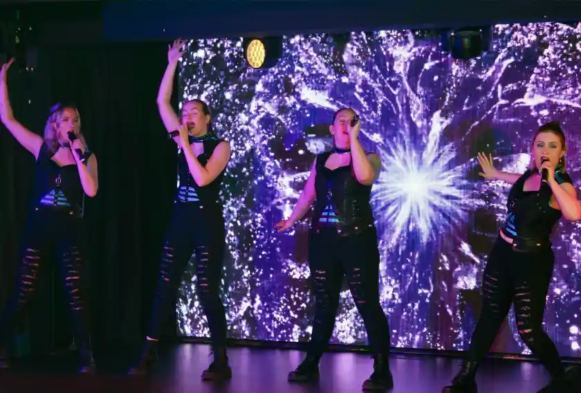 Four female performers in black outfits sing and dance on a stage with a colorful, sparkling background. They hold microphones and pose energetically, showcasing their performance.
