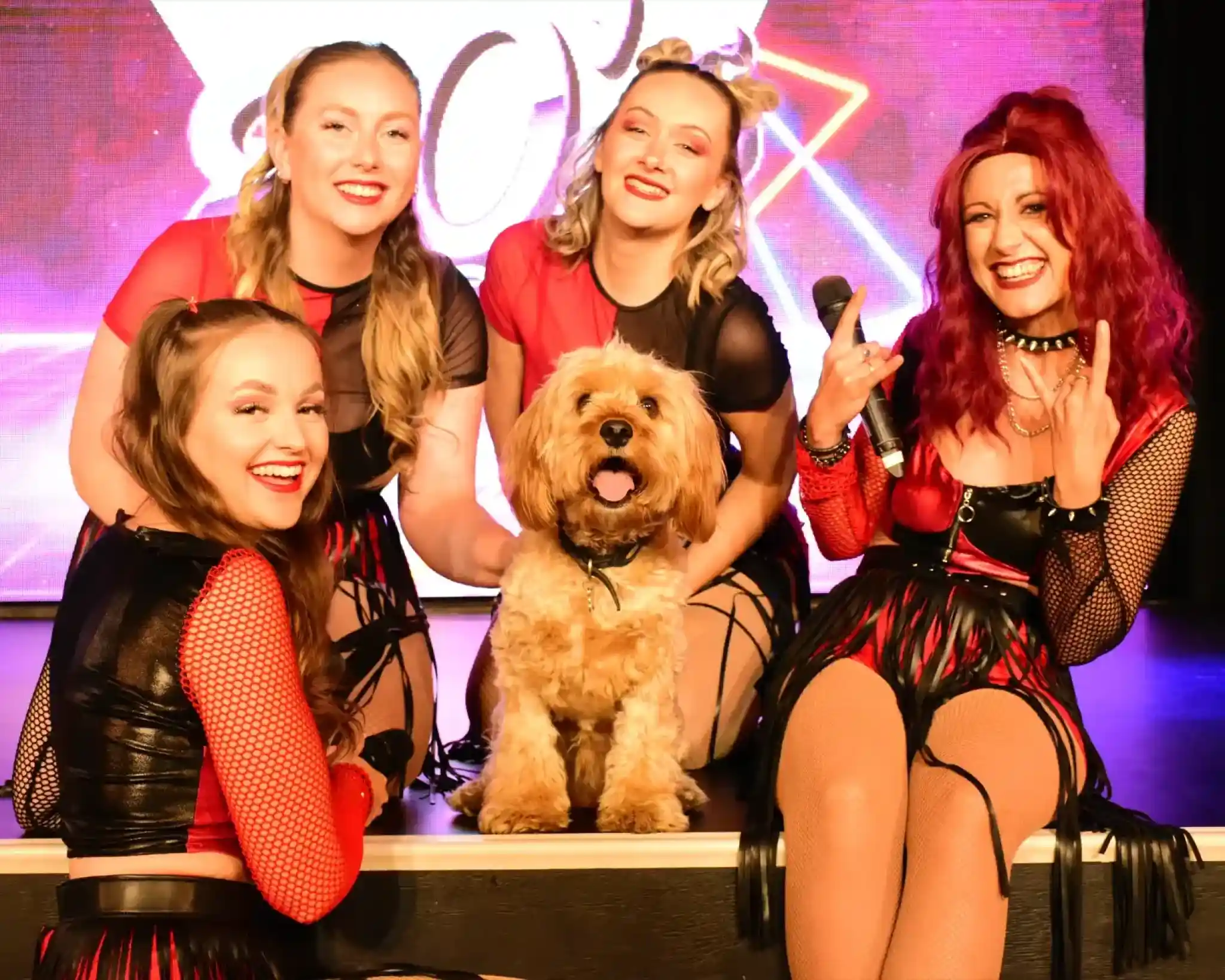 Five performers with vibrant outfits pose together on stage, smiling and making playful gestures. A fluffy dog sits in the center, adding charm to the group. Colorful stage lights are visible in the background.