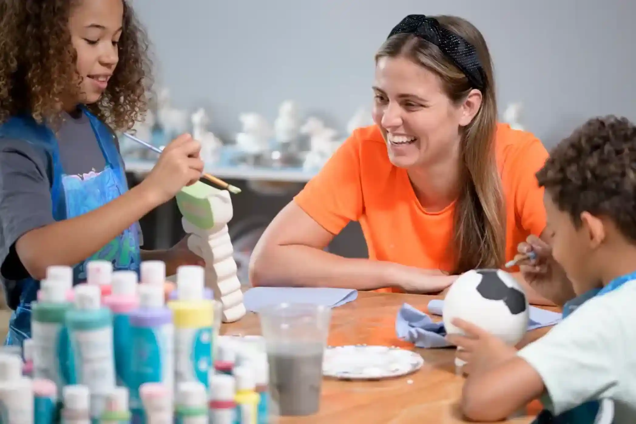 Two children are painting clay figures at a table, while an adult woman watches and smiles. The table is cluttered with paint bottles, brushes, and a paint palette.