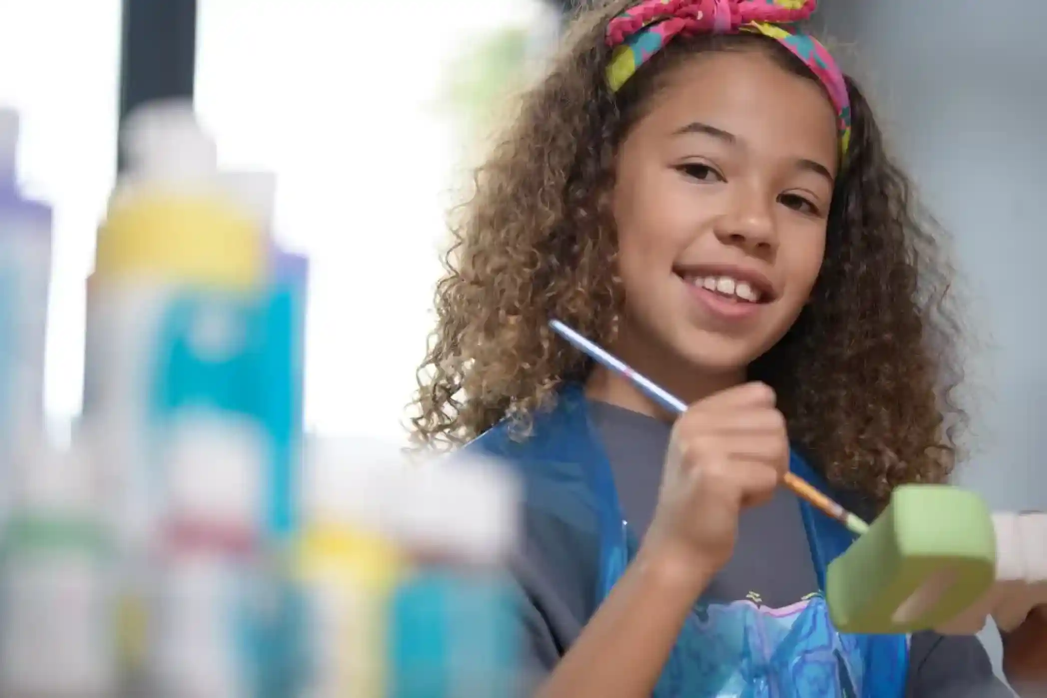 A young girl with curly hair and a colorful headband smiles while holding a paintbrush and working on a green craft project. Bottles of paint are blurred in the background.