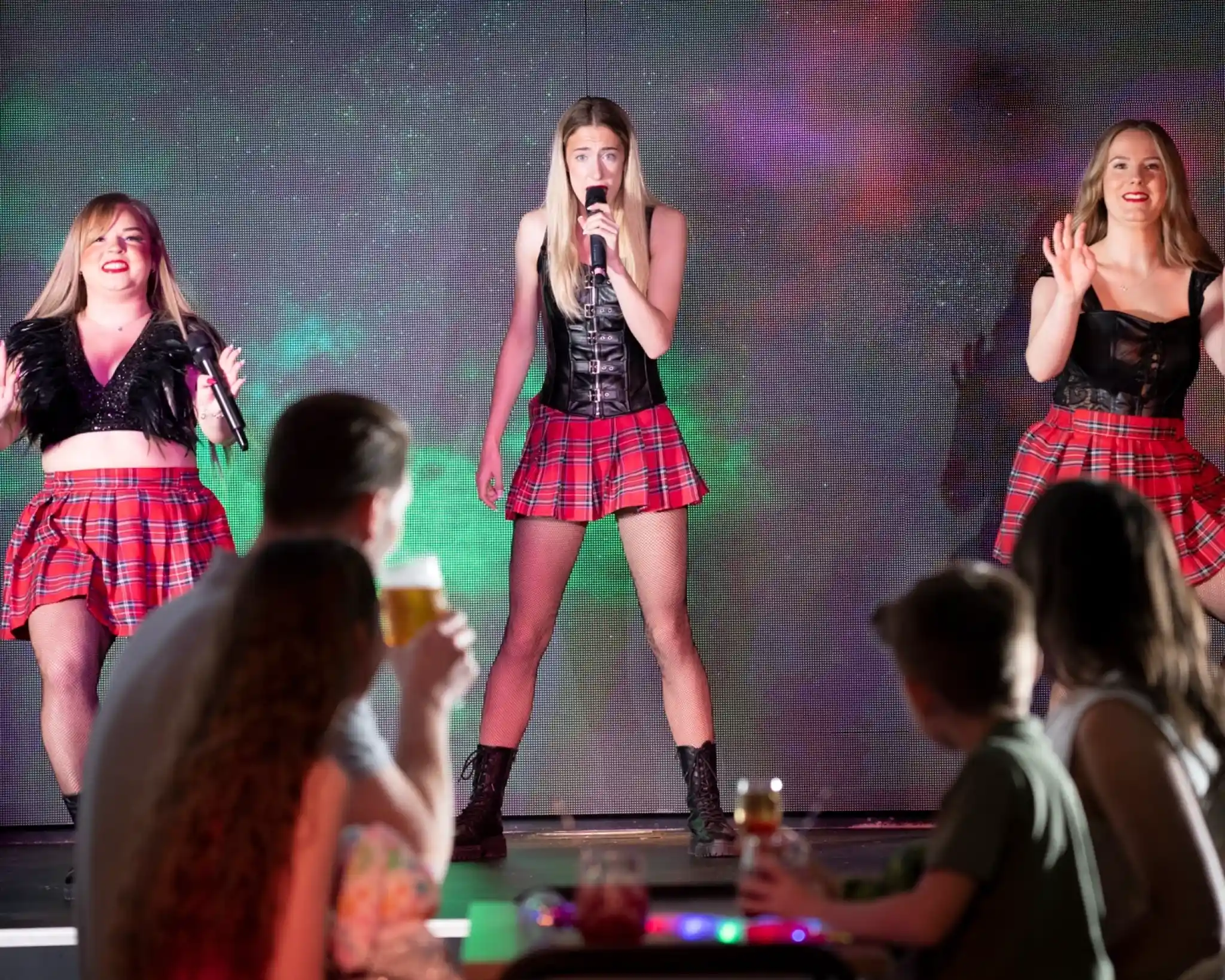 The image shows three female performers on stage, all wearing coordinated outfits consisting of red tartan skirts and black tops (with leather or lace details). The central performer is holding a microphone and singing, while the two on either side are dancing and also appear to be singing or harmonizing. Their confident poses and stage presence suggest a choreographed show, likely musical or dance-based.

In the foreground, audience members—including adults and children—are seated and watching the performance. Some are holding drinks, indicating a relaxed, possibly family-friendly evening event at a holiday venue or resort. There's a vibrant LED screen in the background adding colorful visual effects to the show.
