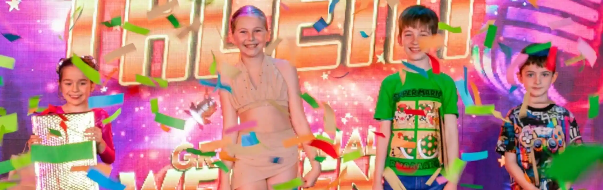 Four young performers celebrate on stage with colorful confetti falling around them. They stand in front of a vibrant backdrop that reads "Shorefield Has Talent." Each holds a prize bag, showcasing their achievements in the talent show.