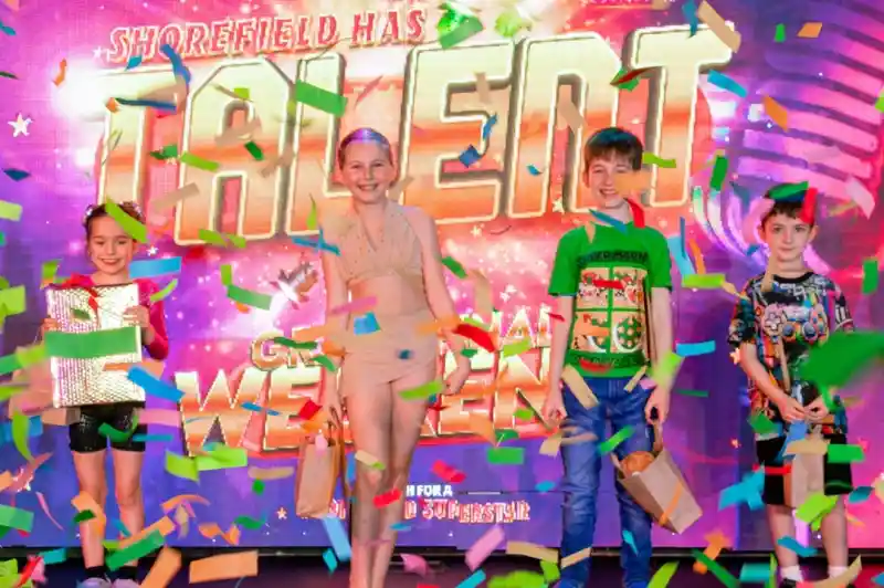 Four young performers celebrate on stage with colorful confetti falling around them. They stand in front of a vibrant backdrop that reads "Shorefield Has Talent." Each holds a prize bag, showcasing their achievements in the talent show.