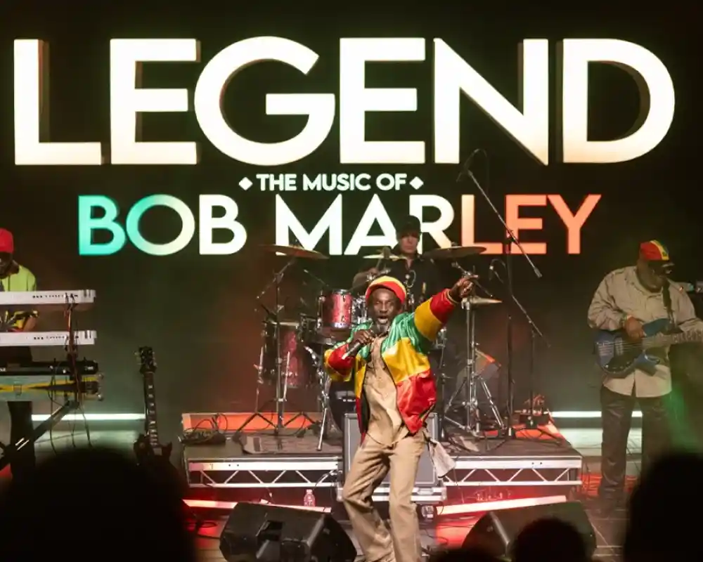 A lively stage performance with a musician in a colorful outfit featuring yellow, green, and red, singing passionately. Behind him, a band plays instruments, including keyboards and drums, with large letters reading "Legend: The Music of Bob Marley" prominently displayed in the background.