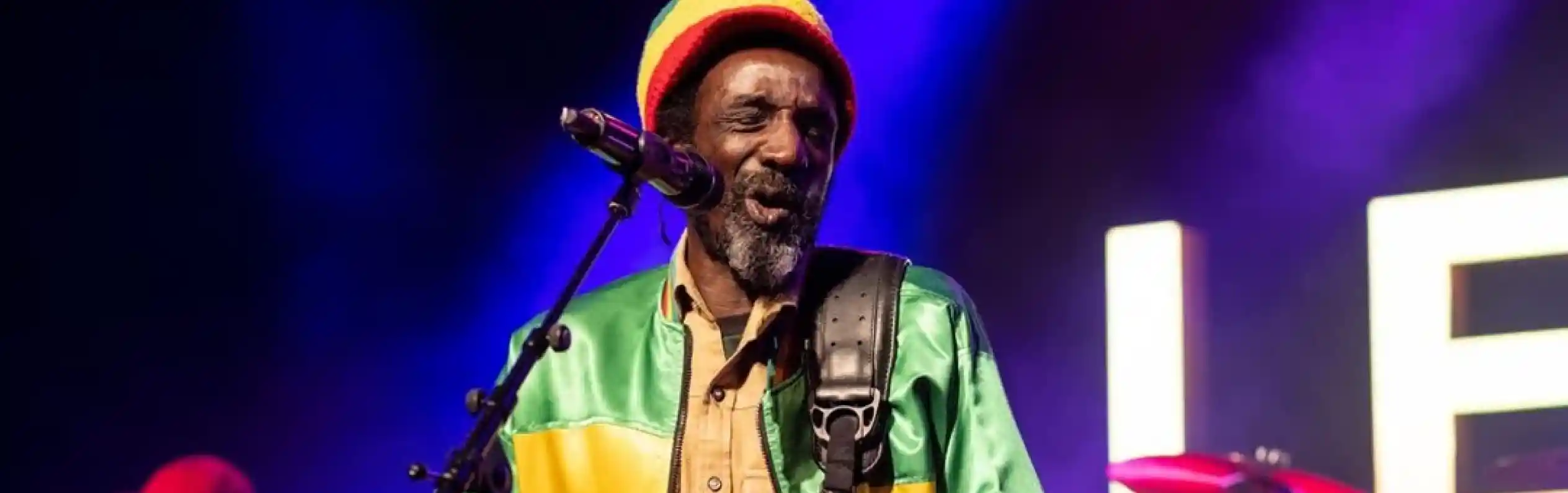 A musician performs on stage, wearing a colorful reggae-style jacket and a crochet beanie. He plays an electric guitar and sings into a microphone, with a keyboardist visible in the background and vibrant concert lights illuminating the scene.