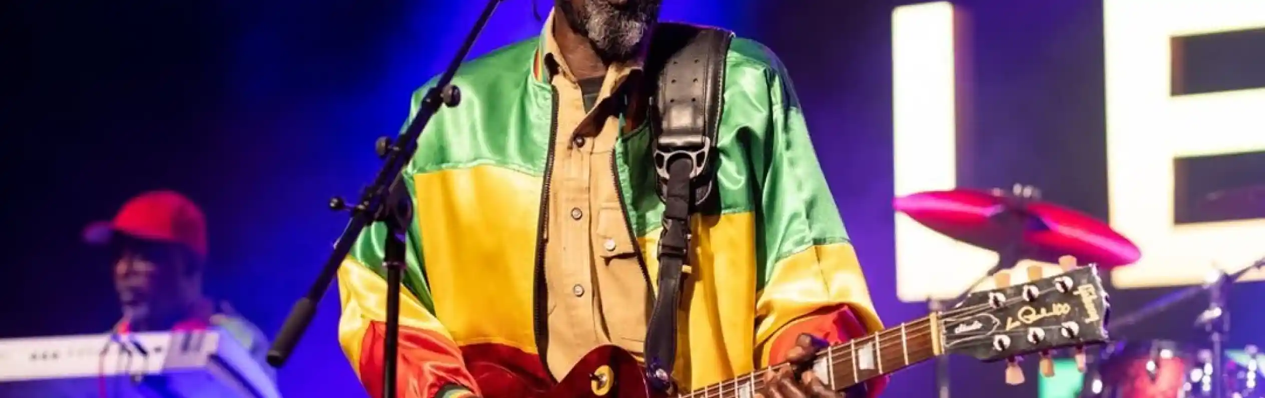 A musician performs on stage, wearing a colorful reggae-style jacket and a crochet beanie. He plays an electric guitar and sings into a microphone, with a keyboardist visible in the background and vibrant concert lights illuminating the scene.