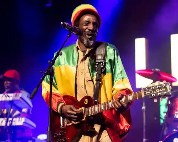 A musician performs on stage, wearing a colorful reggae-style jacket and a crochet beanie. He plays an electric guitar and sings into a microphone, with a keyboardist visible in the background and vibrant concert lights illuminating the scene.