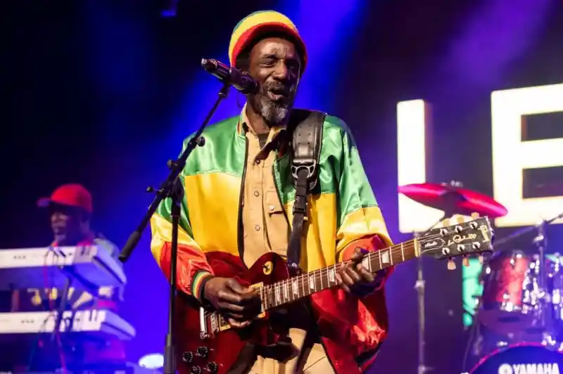A musician performs on stage, wearing a colorful reggae-style jacket and a crochet beanie. He plays an electric guitar and sings into a microphone, with a keyboardist visible in the background and vibrant concert lights illuminating the scene.