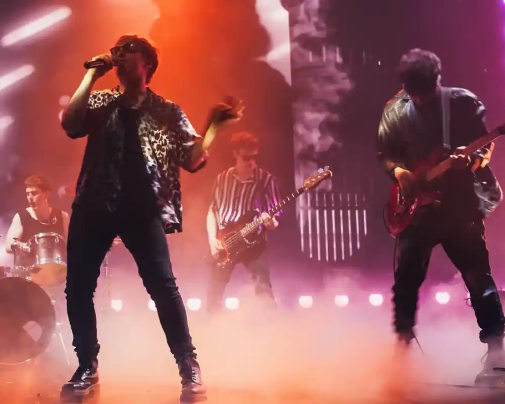 A band performs on stage amidst colorful lights and smoke. The lead singer, wearing sunglasses and a patterned shirt, holds a microphone. A guitarist and bassist accompany him, while a drummer is visible in the background.