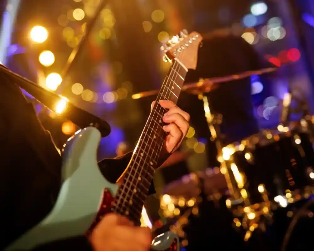 A musician plays an electric guitar with a blue body while a drummer is visible in the background, illuminated by colourful stage lights.
