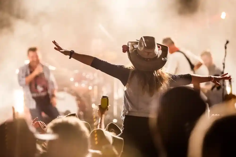 A concert scene with a crowd enjoying the performance. A woman in a hat stands with her arms outstretched, facing the stage, while a musician performs in the background, surrounded by stage lights and smoke.