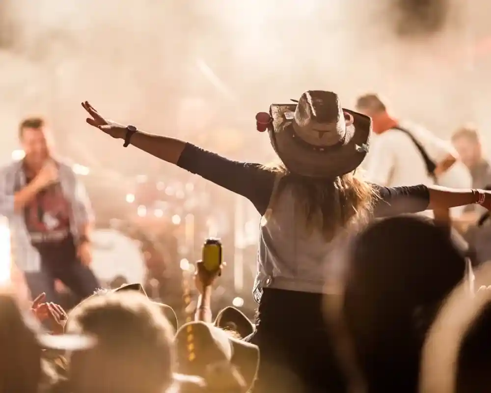 A concert scene with a crowd enjoying the performance. A woman in a hat stands with her arms outstretched, facing the stage, while a musician performs in the background, surrounded by stage lights and smoke.