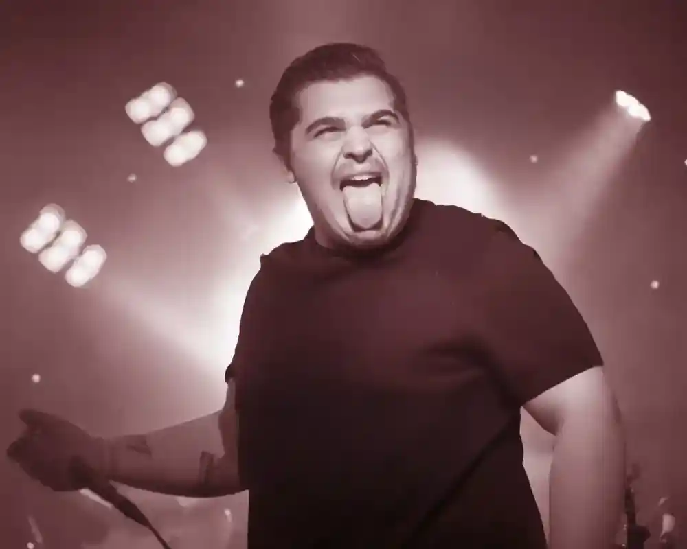 A male singer with a short haircut makes a playful expression, sticking out his tongue while performing on stage. Bright stage lights create a dramatic atmosphere in the background.