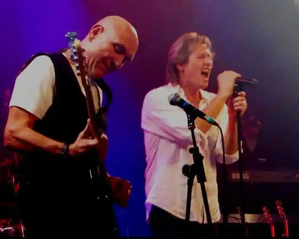 Two male musicians perform on stage. One plays an electric guitar with a focused expression, while the other passionately sings into a microphone, both surrounded by colorful stage lighting.