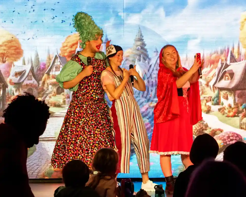 Three performers on stage interact with the audience. Two are dressed in colorful costumes; one wears a large green wig and a floral dress, while the other wears a red cape. The third performer, in striped overalls, holds a microphone and takes a selfie. A vibrant, animated backdrop features a whimsical landscape. Children are visible in the foreground watching the show.