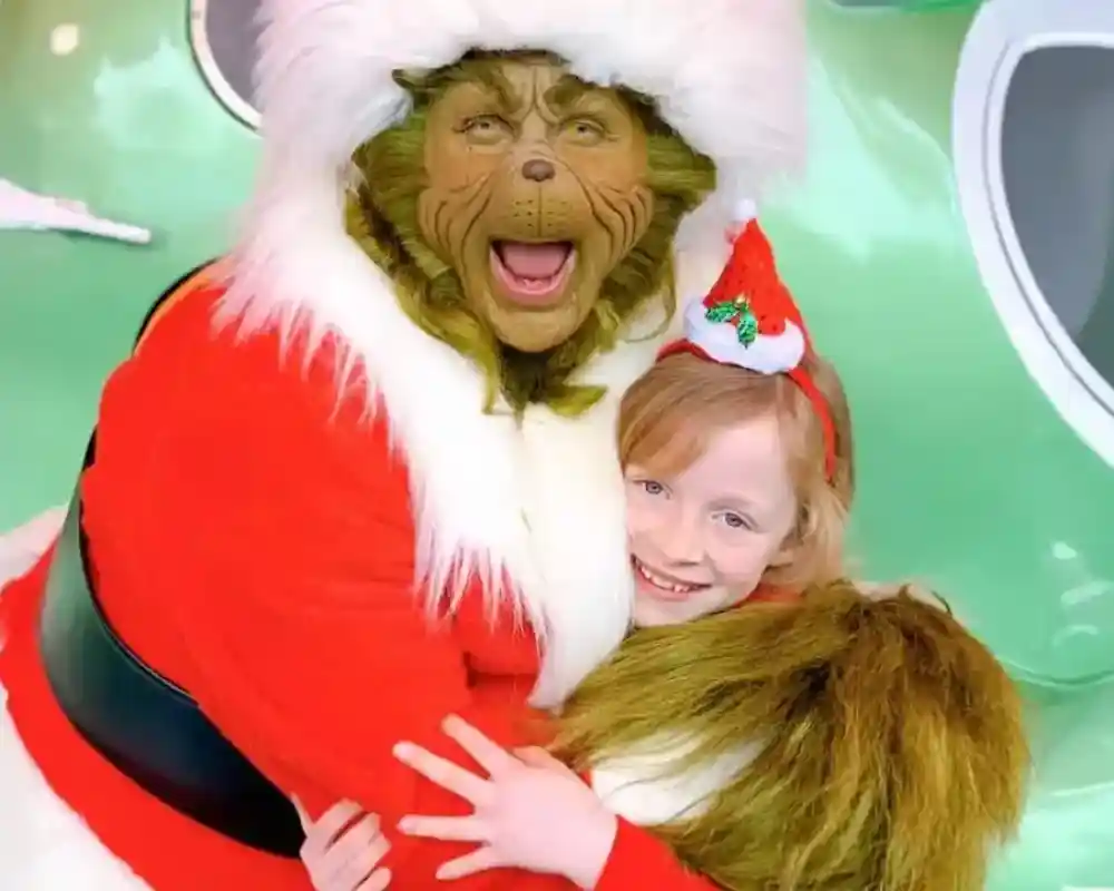 A person in a Santa costume with a Grinch mask joyfully hugs a young girl wearing a festive headband. They are surrounded by a colorful holiday-themed background.