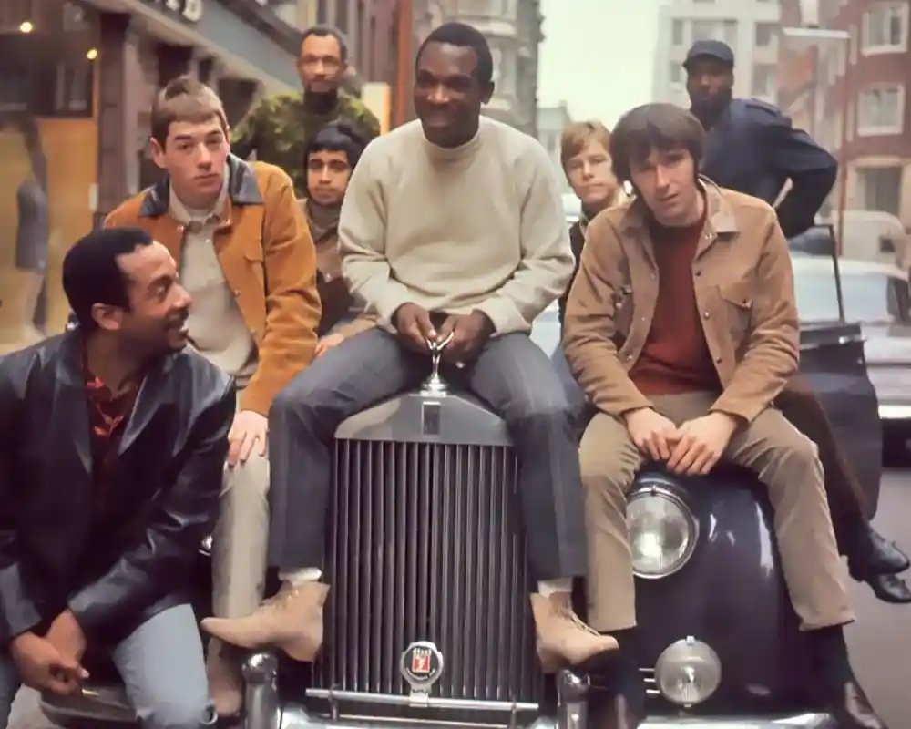 A group of six men poses on and around a vintage car in an urban setting. They display a mix of casual clothing styles typical of the 1960s, with some smiling and interacting with each other. The city backdrop includes buildings and street life.