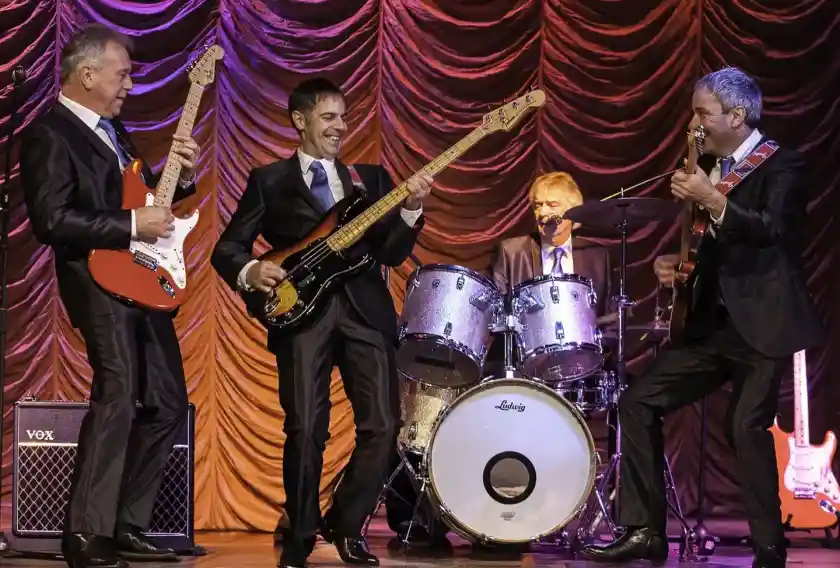 A band of four musicians performing on stage, dressed in suits. Two guitarists play electric guitars while a bassist stands in the center, smiling. A drummer sits behind a drum kit, contributing to the lively atmosphere with colorful stage lighting in the background.