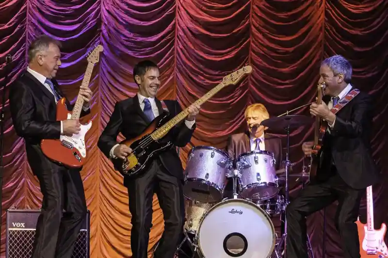 A band of four musicians performing on stage, dressed in suits. Two guitarists play electric guitars while a bassist stands in the center, smiling. A drummer sits behind a drum kit, contributing to the lively atmosphere with colorful stage lighting in the background.