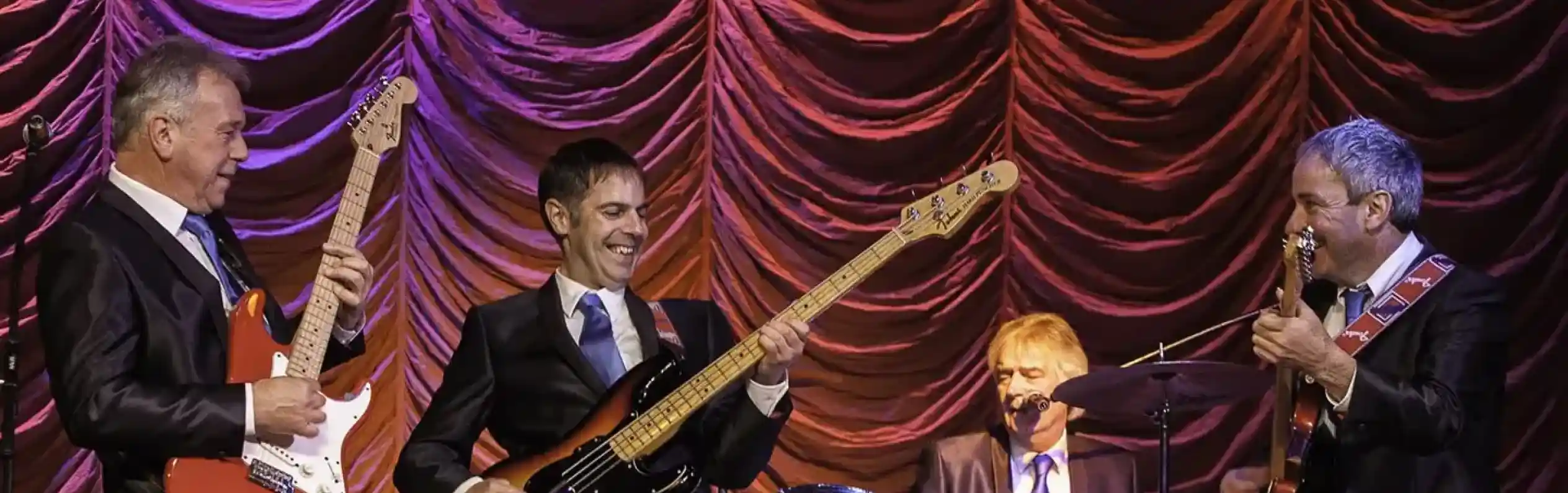 A band of four musicians performing on stage, dressed in suits. Two guitarists play electric guitars while a bassist stands in the center, smiling. A drummer sits behind a drum kit, contributing to the lively atmosphere with colorful stage lighting in the background.