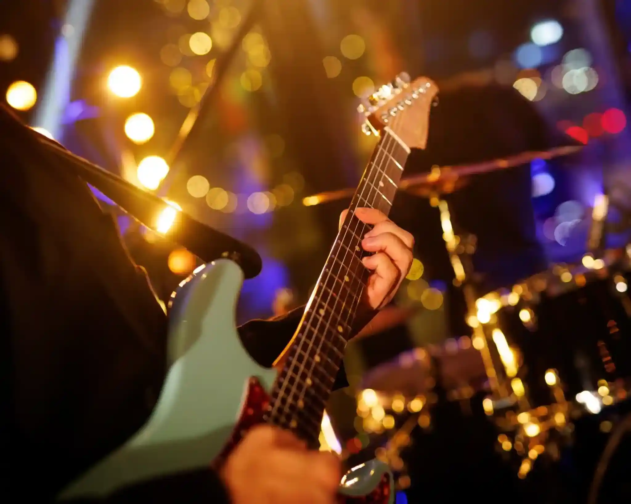 A close-up of a musician's hand playing a light blue electric guitar, with blurred colorful stage lights and a drum set in the background.