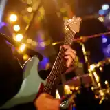 A close-up of a musician's hand playing a light blue electric guitar, with blurred colorful stage lights and a drum set in the background.