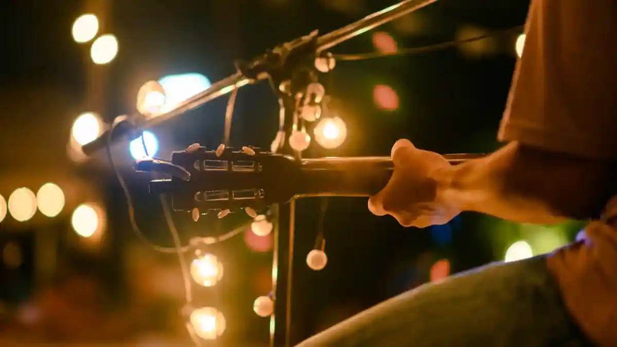 A band of four musicians performing on stage, dressed in suits. Two guitarists play electric guitars while a bassist stands in the center, smiling. A drummer sits behind a drum kit, contributing to the lively atmosphere with colorful stage lighting in the background.