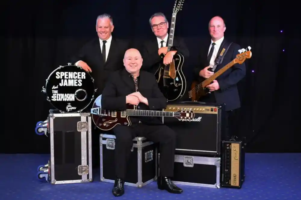 A group of four male musicians dressed in black suits poses together. The man in front sits on a case holding an electric guitar, while the others stand behind him with various instruments, including a bass guitar and a drum. They are arranged against a dark backdrop with subtle lighting.
