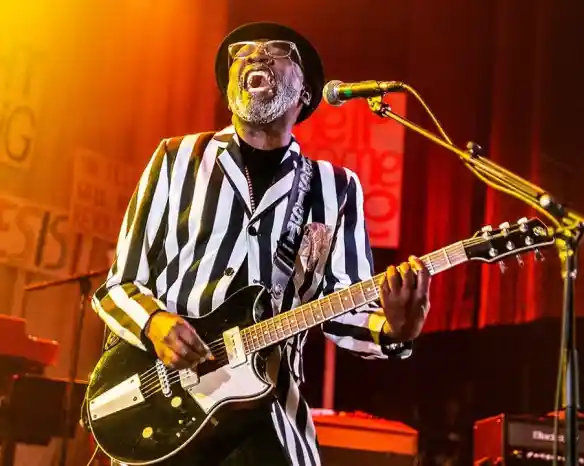 A musician wearing a striped suit and hat plays an electric guitar while passionately singing on stage. Bright stage lights create an energetic atmosphere, and colorful signs are visible in the background.