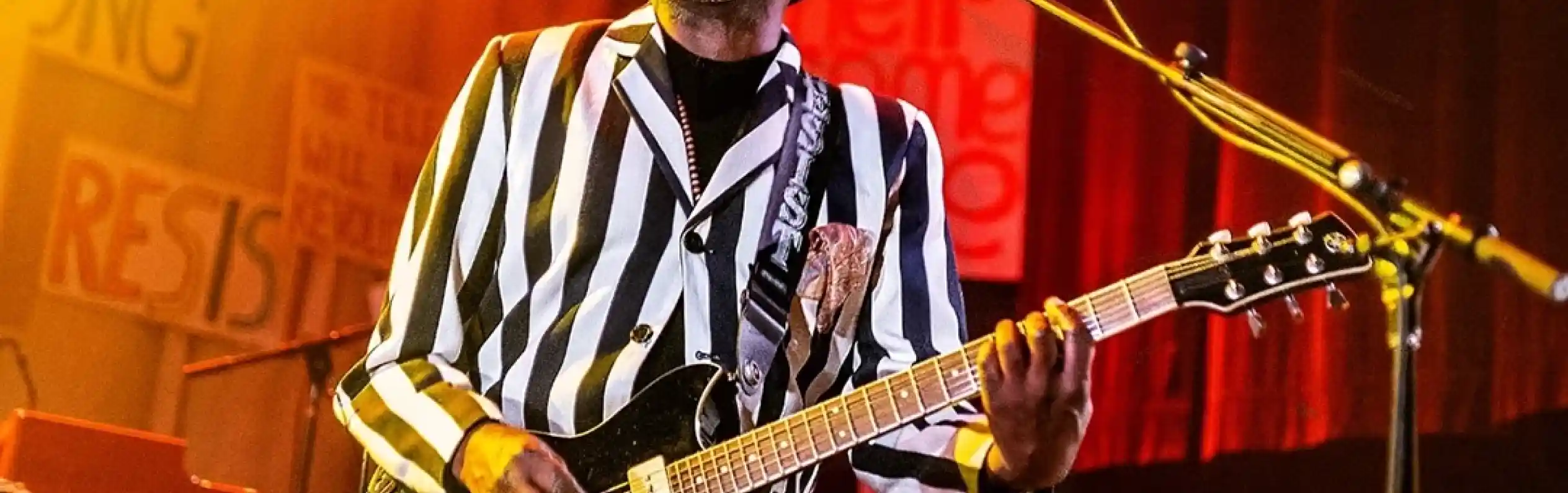A musician wearing a striped suit and hat plays an electric guitar while passionately singing on stage. Bright stage lights create an energetic atmosphere, and colorful signs are visible in the background.
