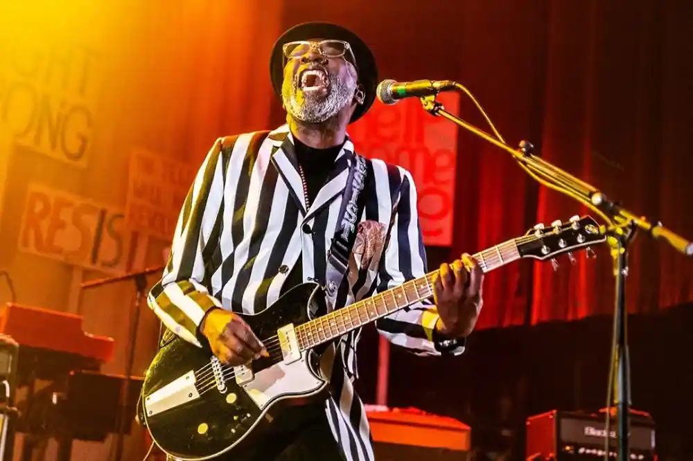 A musician wearing a striped suit and hat plays an electric guitar while passionately singing on stage. Bright stage lights create an energetic atmosphere, and colorful signs are visible in the background.