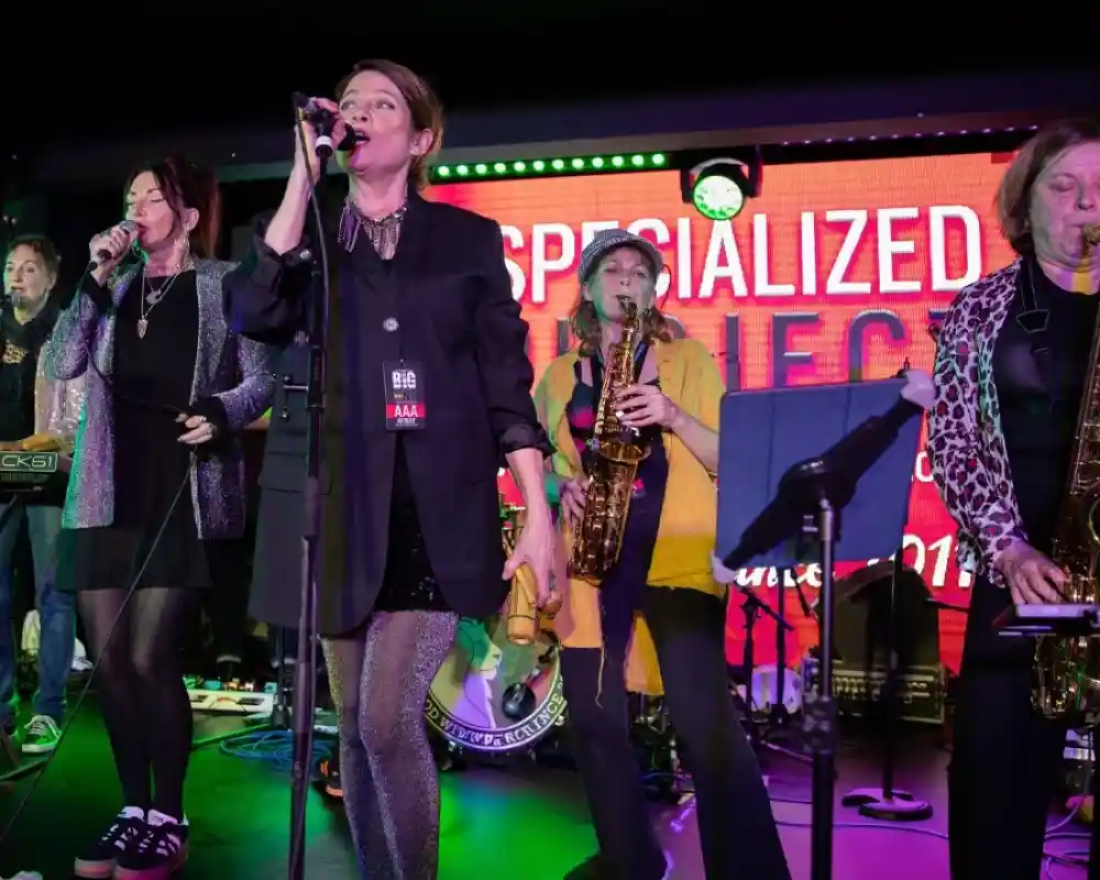 A group of six musicians performing on stage, with one vocalist at the front singing into a microphone. The musicians are dressed in various styles and playing different instruments, including keyboard and saxophones. Colorful stage lights and a vibrant backdrop add to the atmosphere.