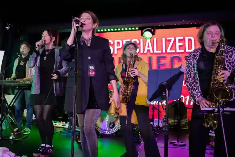 A group of six musicians performing on stage, with one vocalist at the front singing into a microphone. The musicians are dressed in various styles and playing different instruments, including keyboard and saxophones. Colorful stage lights and a vibrant backdrop add to the atmosphere.