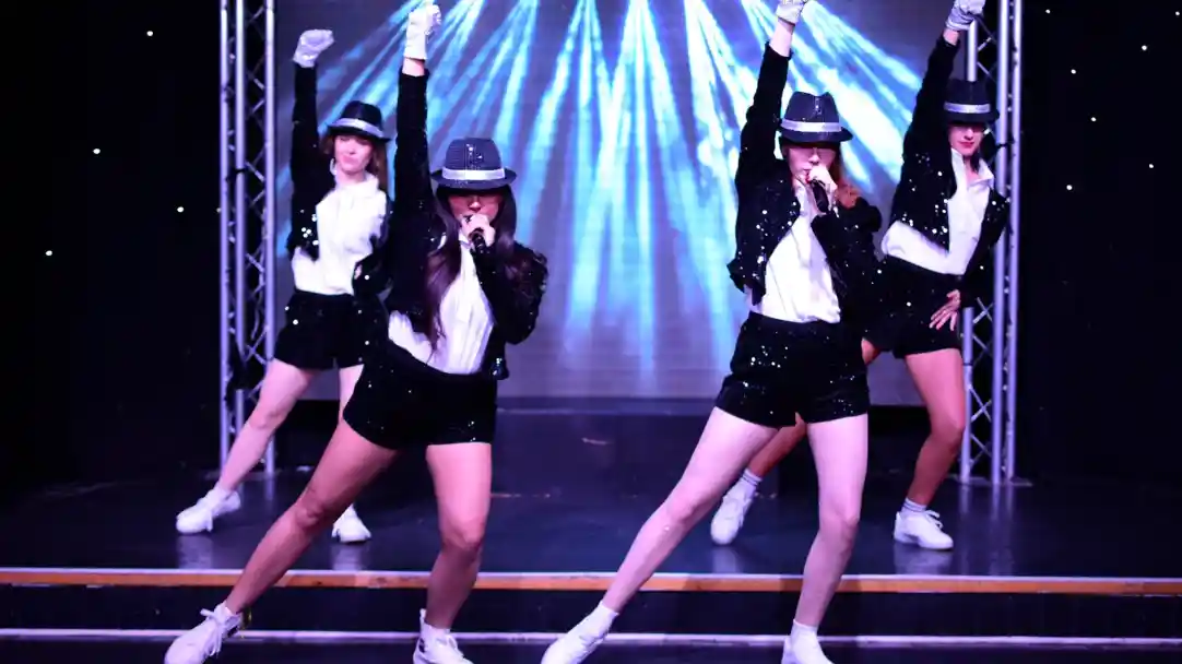 A group of four female dancers perform on stage, dressed in black and white outfits with hats. They strike dynamic poses, one holding a microphone, while colorful light beams illuminate the background.