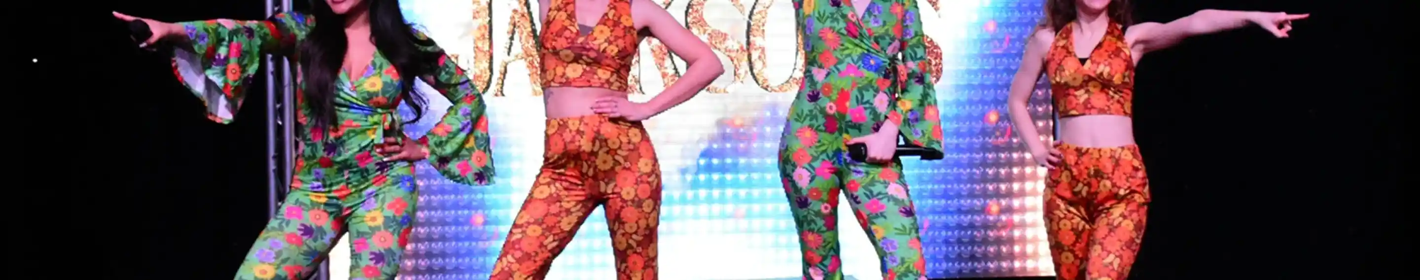 Four dancers perform on stage, wearing colorful, flower-patterned outfits with flared pants and crop tops. They strike dynamic poses with arms raised, set against a vibrant, illuminated background displaying the word "JAMSONS."