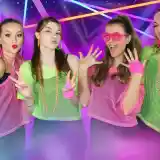 Four young women pose together, wearing colorful neon mesh tops and accessories. They have playful expressions and are surrounded by a vibrant, retro-themed background with geometric patterns and neon lights.