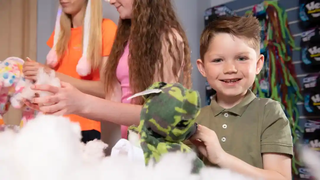 A smiling young boy holds a green camo stuffed animal while two girls in the background work on crafts. The setting appears bright and playful, with fluffy stuffing around them.