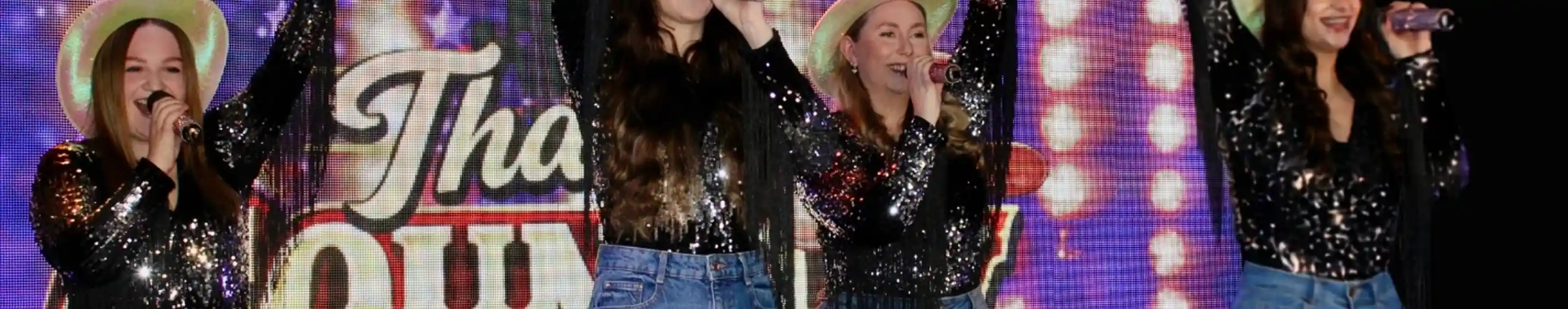 Four performers in sequined tops and denim shorts sing together on stage. They wear cowboy hats and raise their arms in celebration, with a colorful, illuminated backdrop featuring the text "That's Country."