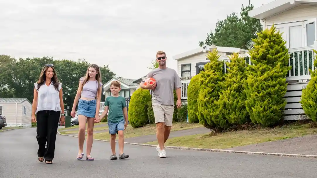 A family walks down a quiet, suburban street, smiling and enjoying their time together. The group includes two adults and two children, one of whom is holding a soccer ball. Lush green shrubs line the pathway, and tidy houses are visible in the background.