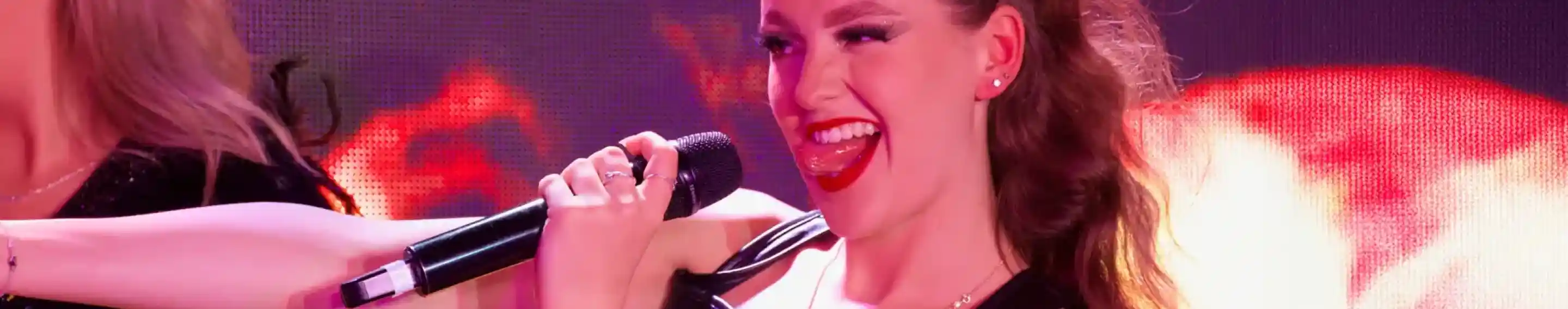 A performer with wavy hair holds a microphone while singing, wearing a black leather outfit. She has a playful expression and bright red lipstick, with a fiery background enhancing the energetic atmosphere.