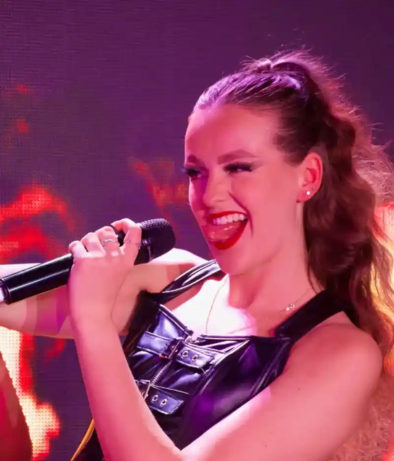 A couple stands on a balcony with a modern home in the background. The man and woman are smiling, dressed casually, with green grass and a landscaped area surrounding the house. The sky is clear and blue.