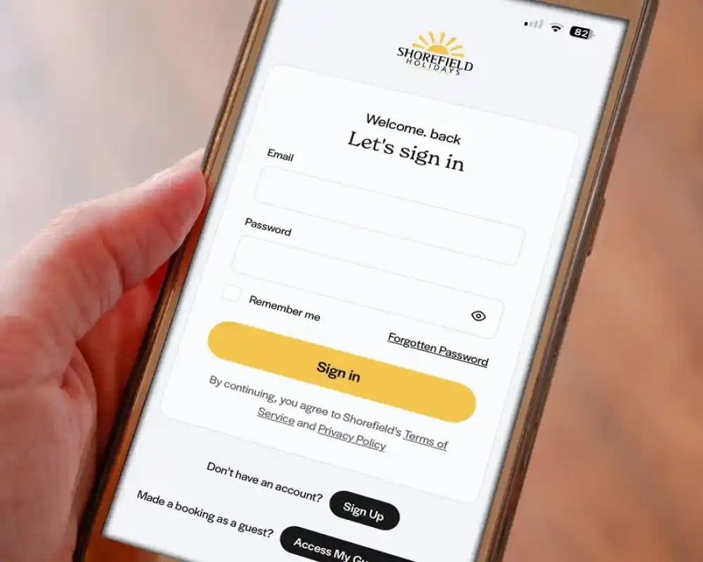 A hand holding a smartphone displaying a login screen for Shorefield Hotels. The screen prompts for email and password input, with options to remember the user and a link for forgotten passwords.