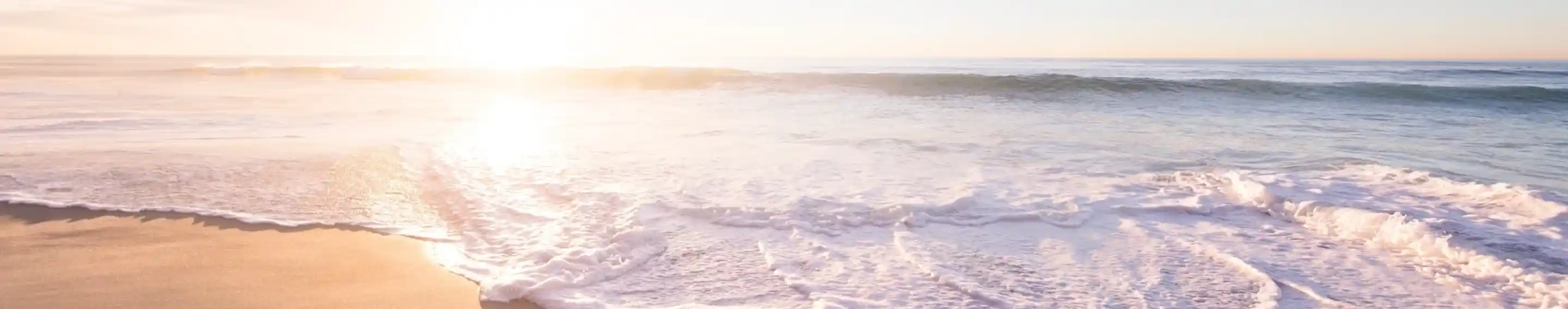 Gentle waves wash onto a sandy beach at sunset, with soft pastel colors in the sky and sunlight reflecting on the water.